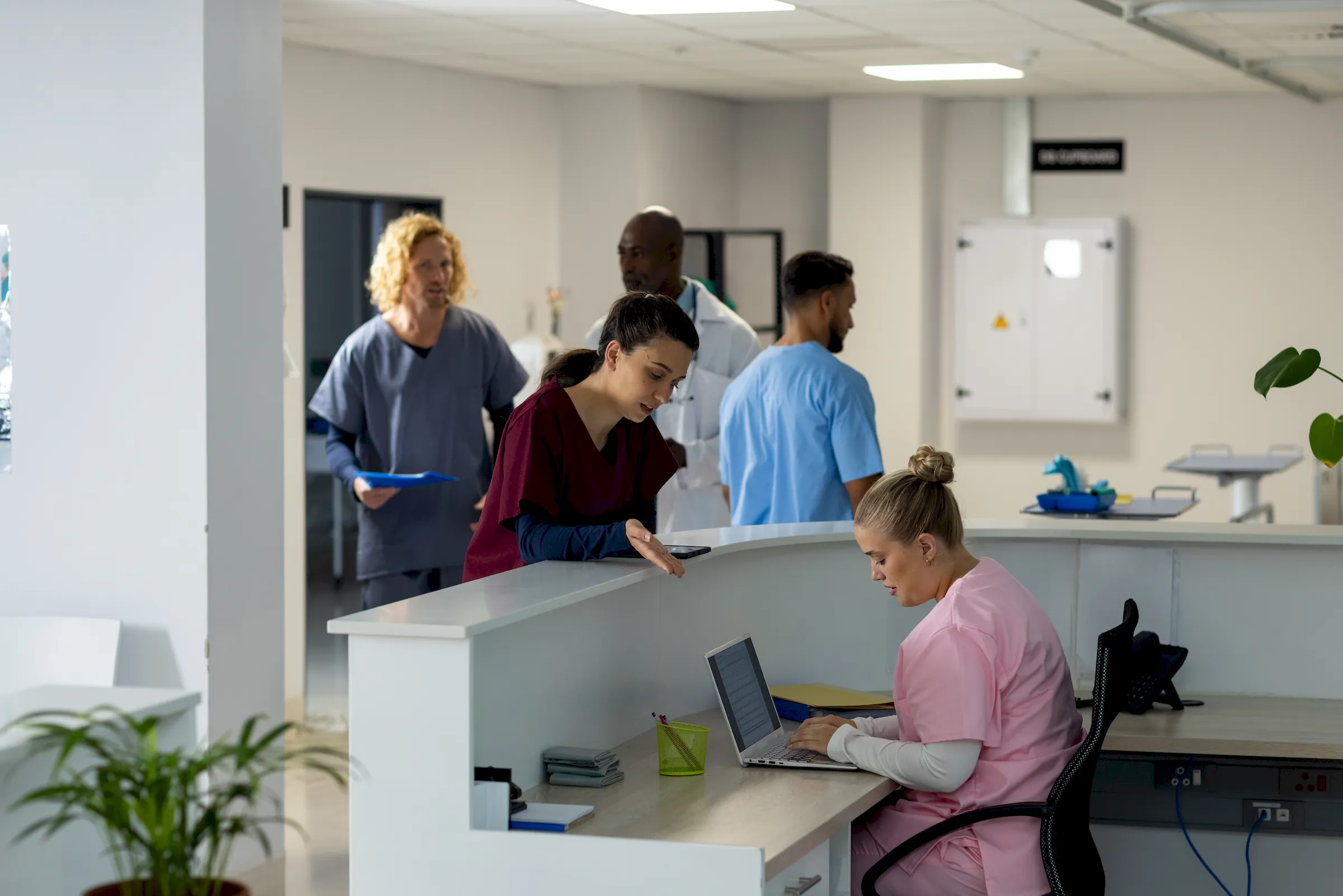 Clinical staff at a hospital nurses' station, one working at a laptop while colleagues confer behind.